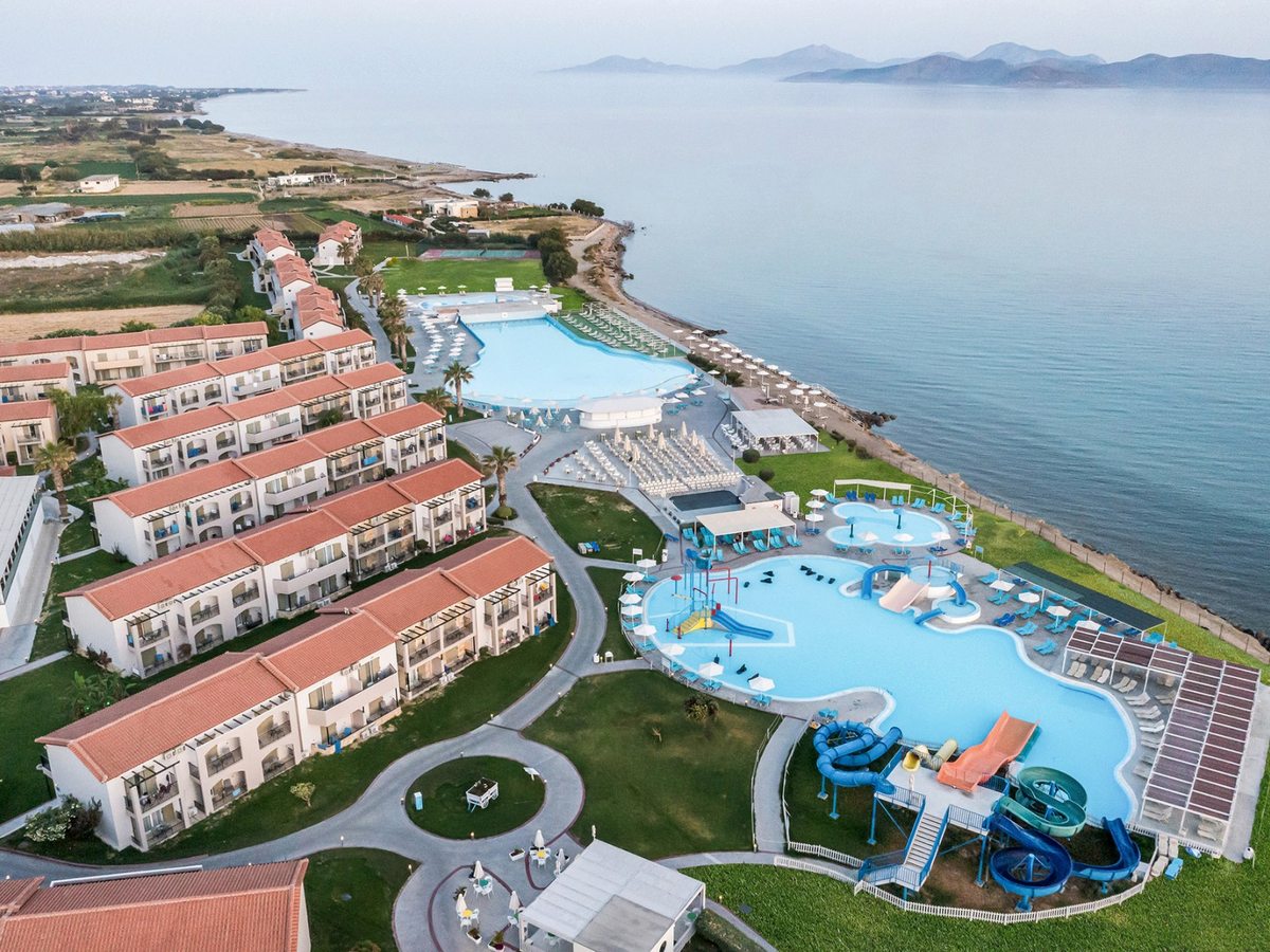 Aerial view of the resort: rows of red-roofed two-storey blocks on lawns, a large freeform pool and the aquapark slides along the shore, with the sea and distant hills beyond.