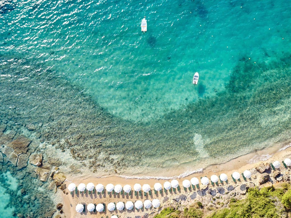 Aerial view of a curve of sand lined with rows of white parasols and sunbeds, with clear shallow water and two small boats.