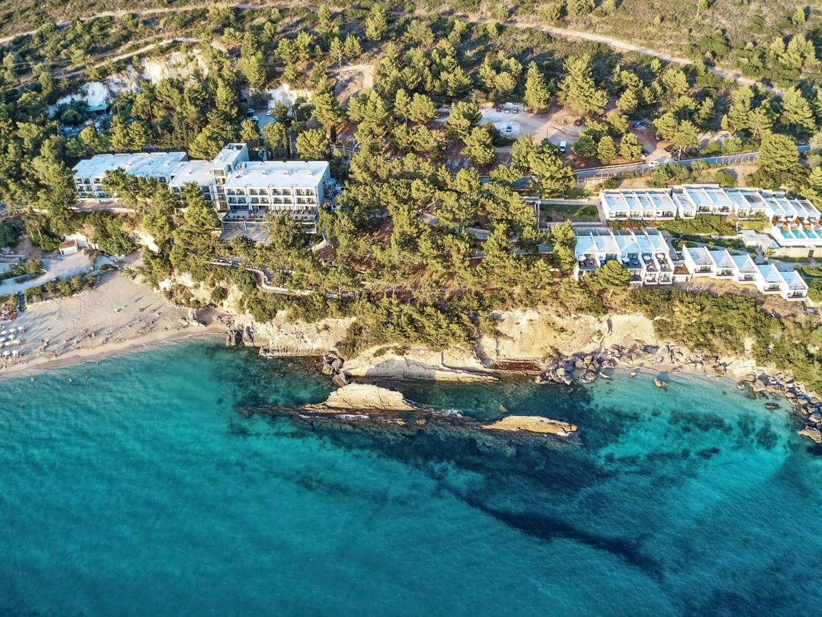 Aerial view of a white hotel building and rows of low white bungalows among pine trees on a headland, above a sandy beach and clear turquoise sea.