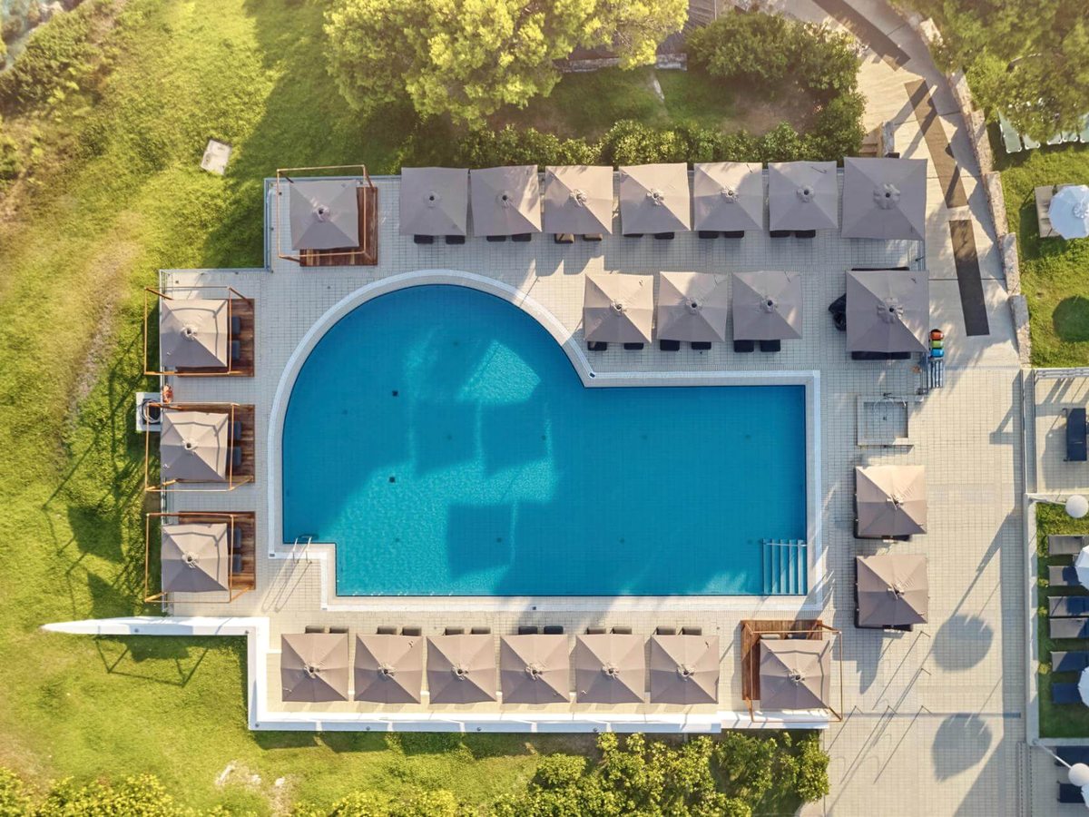 Overhead view of the pool with rows of square parasols and sunbeds on the paved terrace around it, and lawns and paths beyond.