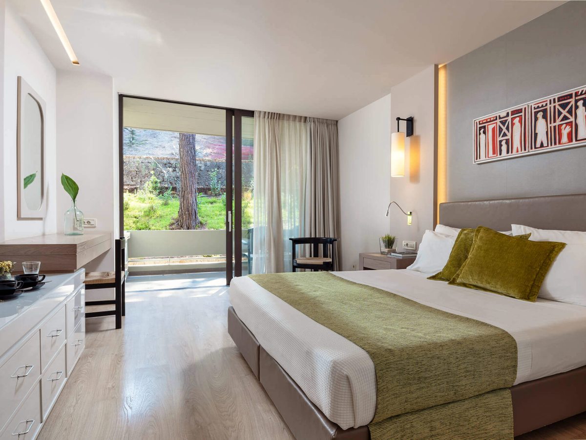 Hotel room with a king bed, a white dresser and a desk, with sliding doors onto a small terrace facing a garden and a scrubby hillside.