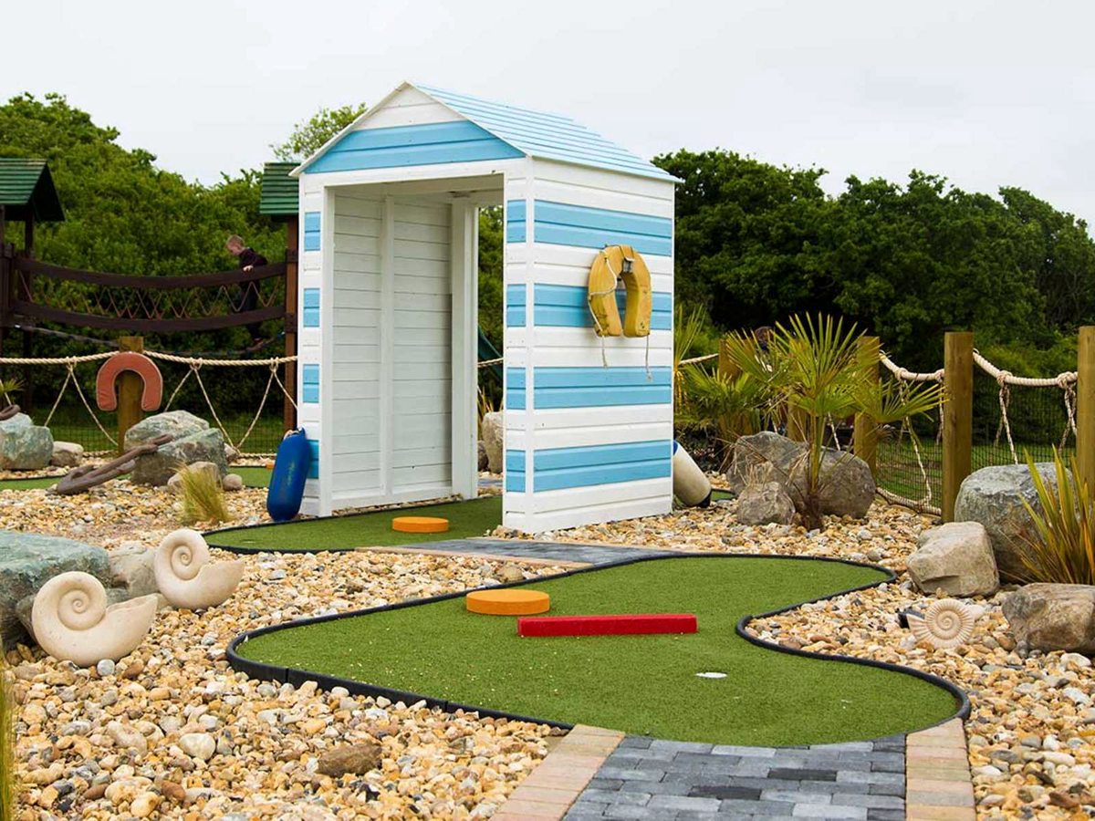 A crazy golf hole with green artificial turf curving around a blue and white striped beach hut, with shell ornaments and pebbles alongside.