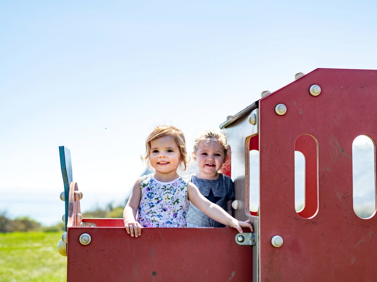 Two toddlers standing in a red wooden play train under a clear sky, with grass and the sea behind.
