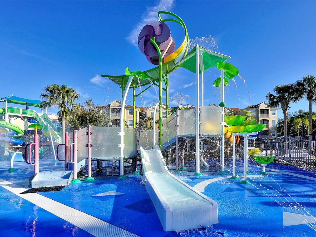 A children's water park with green and yellow slides, a tall water-play tower and a blue splash-pad area among palm trees.