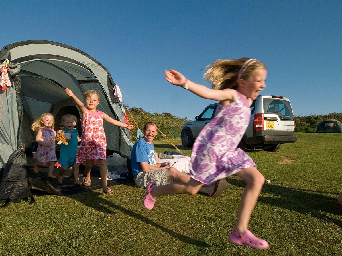 Three young children running and playing outside a large grey family tent on a grass pitch, with a car parked alongside and a man sitting in the tent doorway.