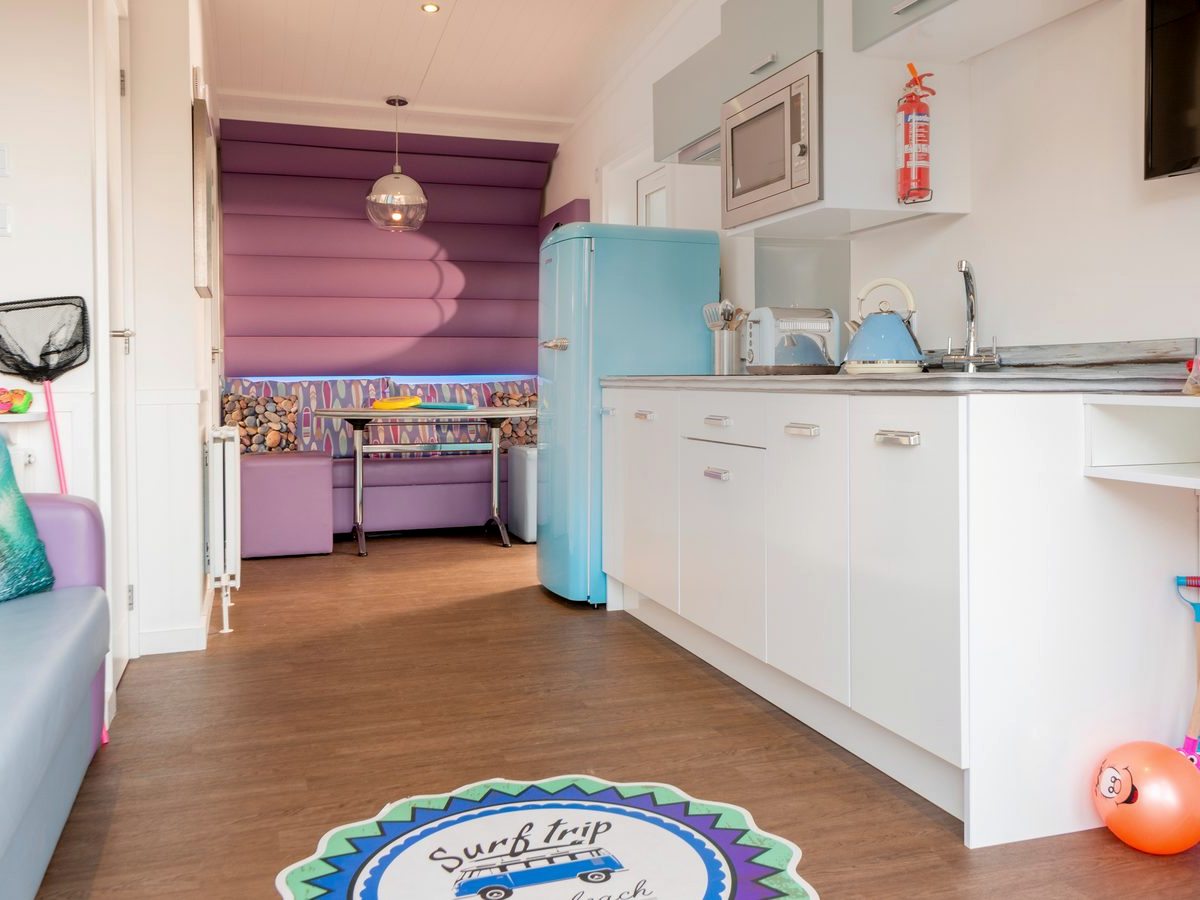Narrow studio interior with a white galley kitchen, a pale blue retro fridge and a purple padded dining nook with a table at the far end.