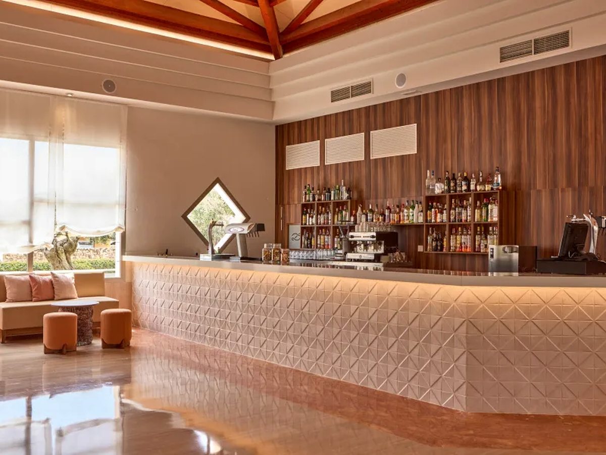 A long bar counter with a backlit textured front, shelves of spirits and a coffee machine behind, and lounge seating to one side under a timber roof.