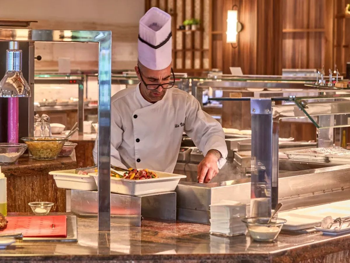 A chef in whites plating grilled vegetables and meat at a live cooking station set into the buffet counter.