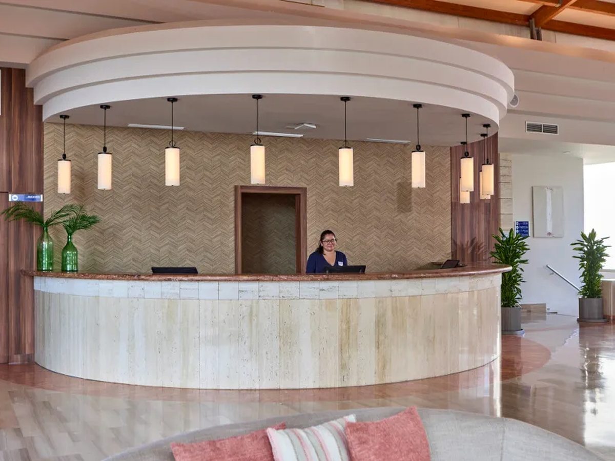A curved stone reception counter under a row of cylindrical pendant lights, a herringbone-panelled wall behind and a member of staff at a computer.
