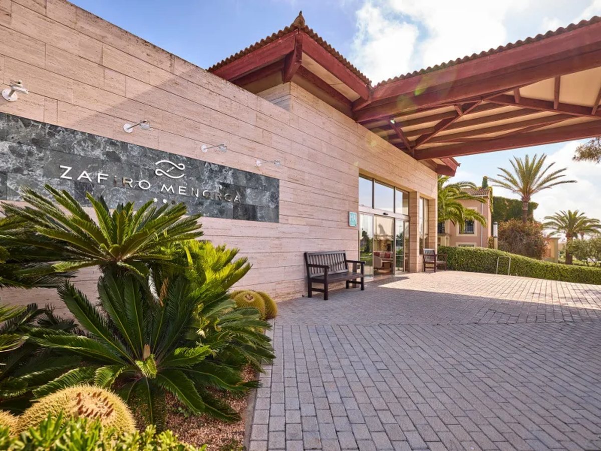 The resort entrance: a stone wall carrying the Zafiro Menorca name and four stars, a timber canopy over a paved driveway, palms and low planting beside a bench.