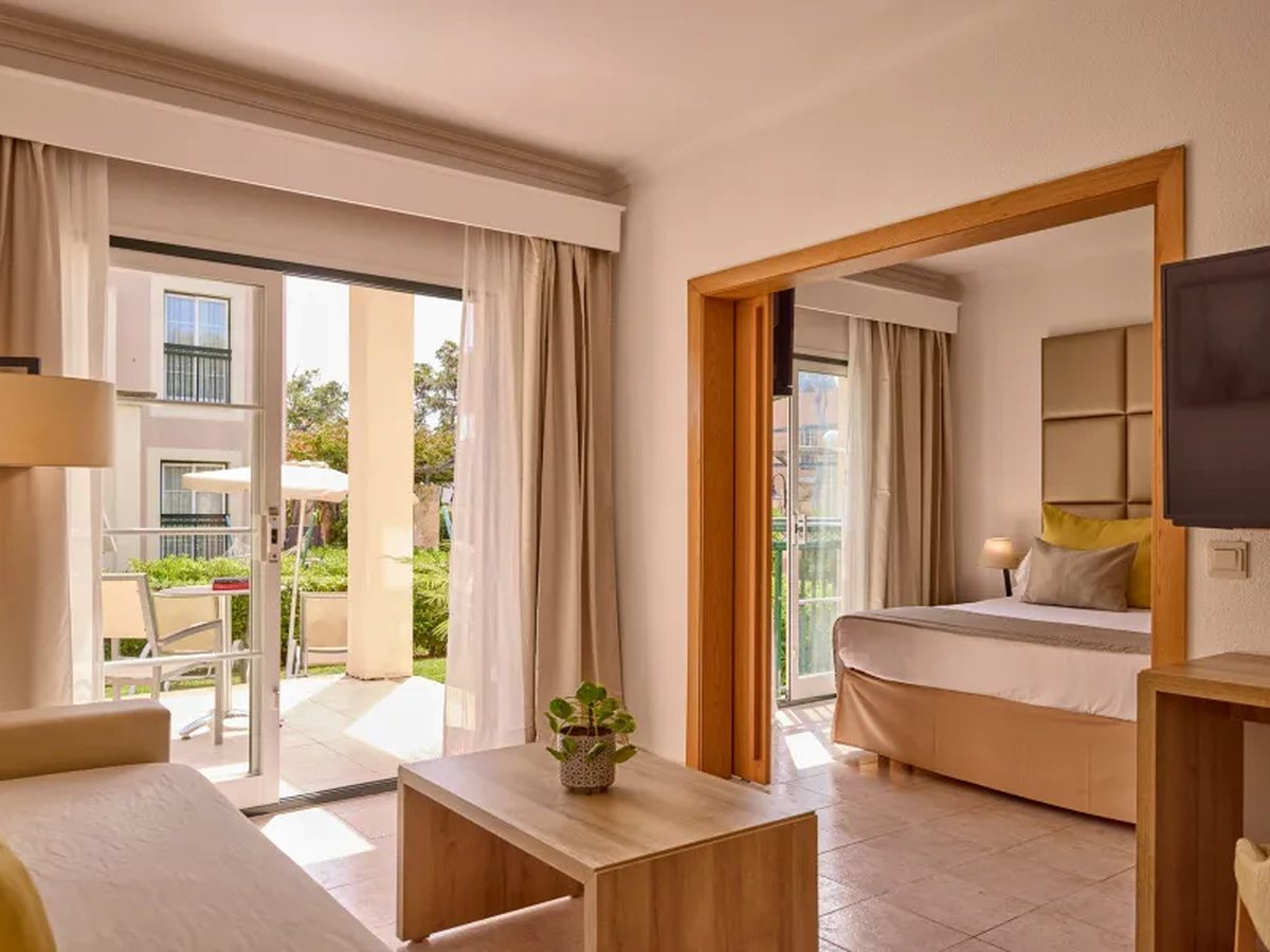 A suite living room with a sofa, low wooden table and wall-mounted TV, sliding doors onto a garden terrace and the bedroom through a wide doorway.
