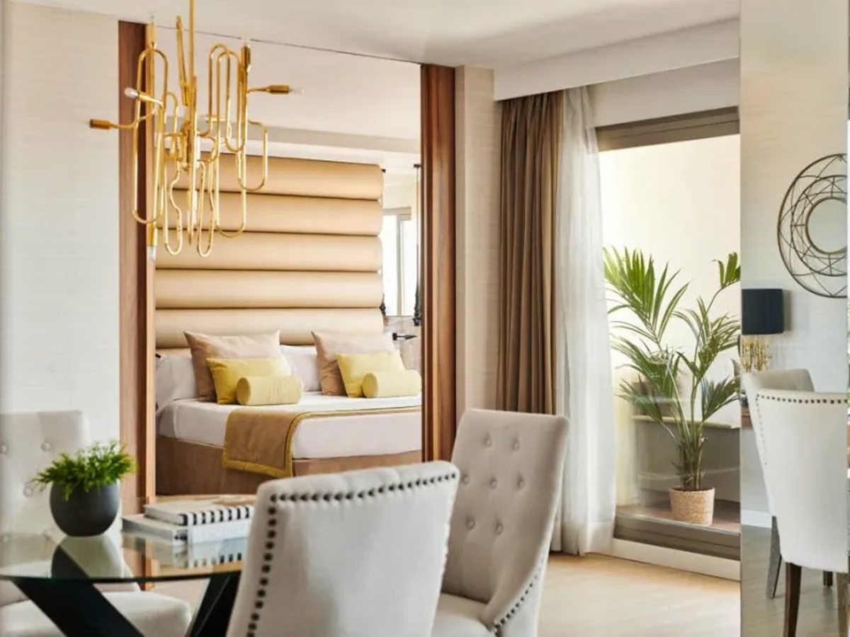 A glass dining table and buttoned chairs in the foreground, with the bed and a gold pendant light through the opening behind.
