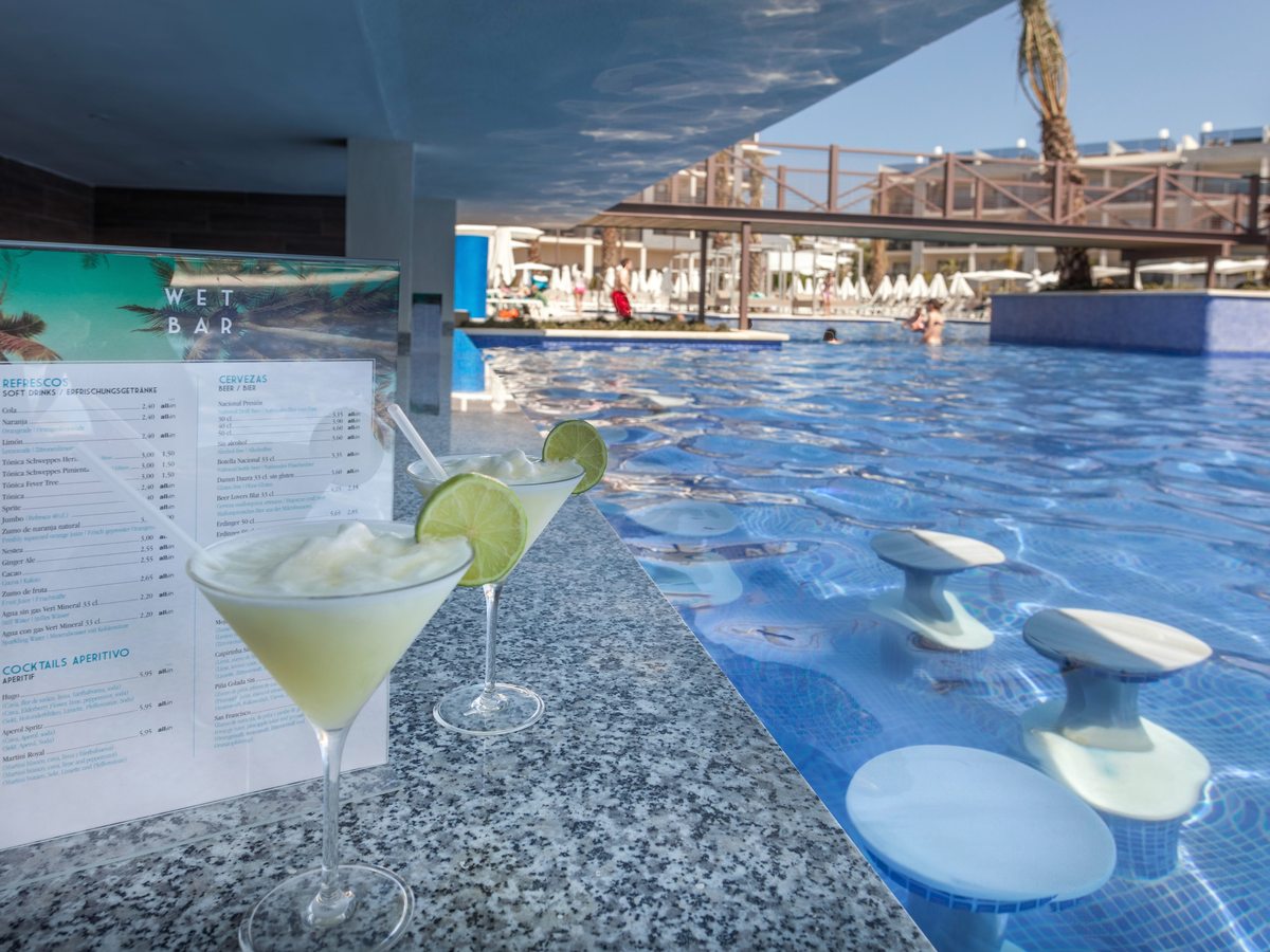 A granite bar top at water level with two frozen cocktails and a drinks menu, and white stools set in the shallow blue pool alongside.