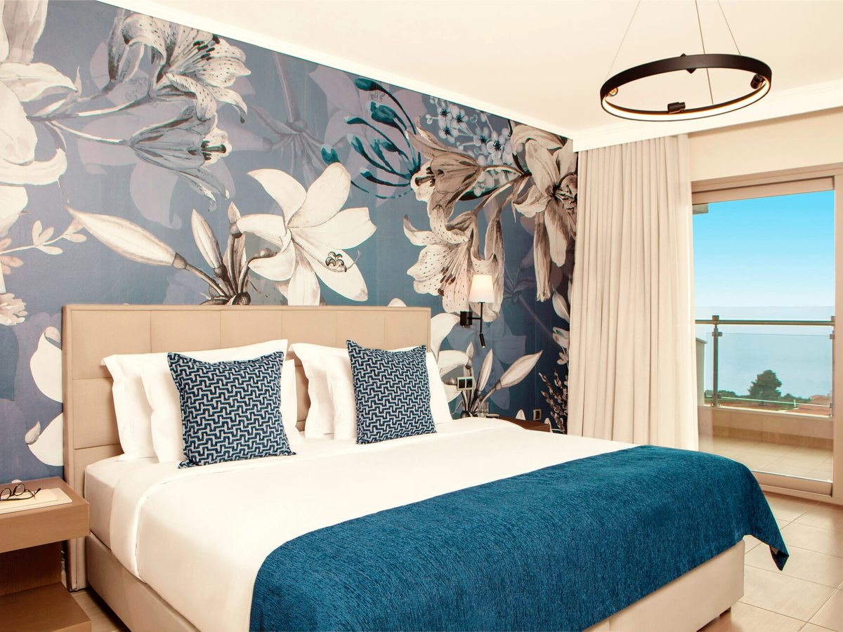A double bed with a teal throw and patterned cushions against a large grey-and-white floral mural, with a balcony door open to the sea.