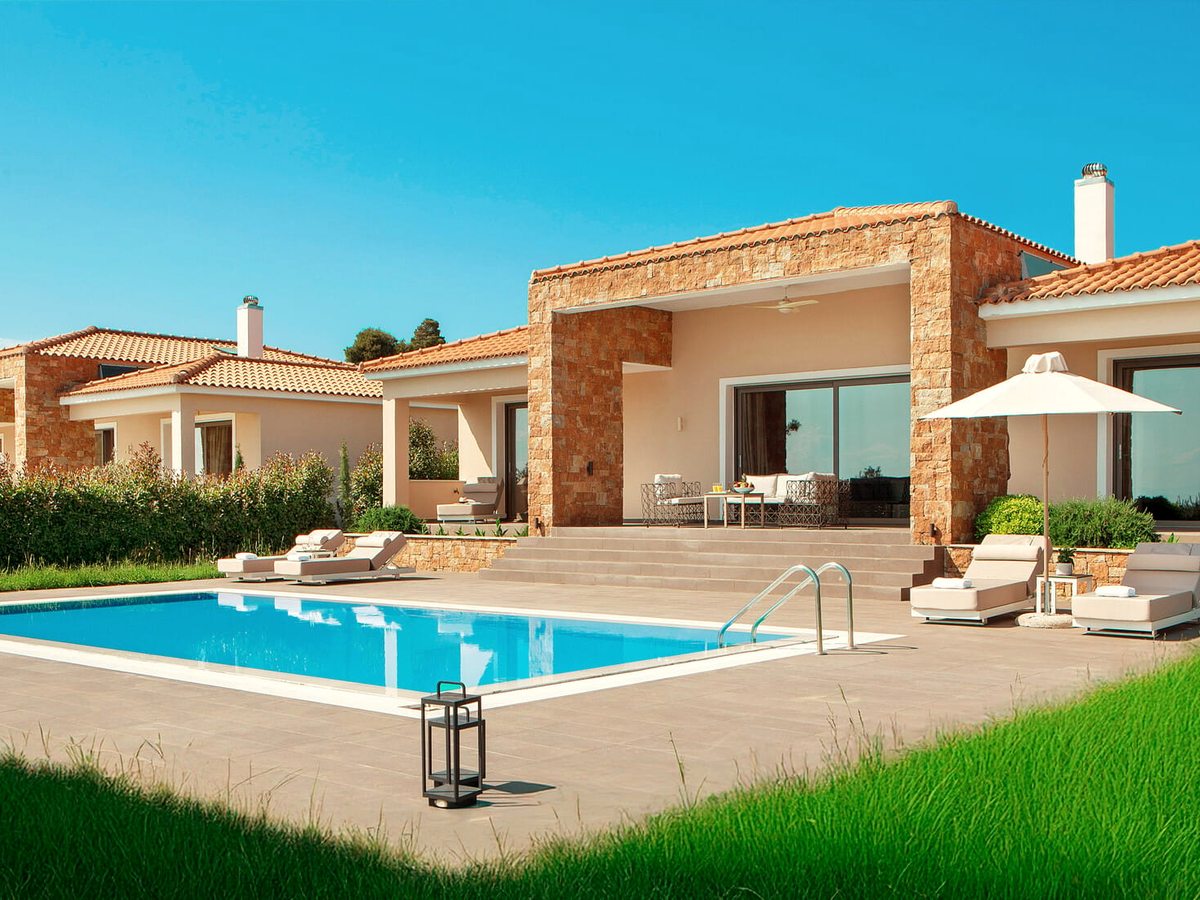 A stone-and-render villa with a terracotta roof beside a rectangular private pool, with sun loungers on the paving and a canvas parasol.