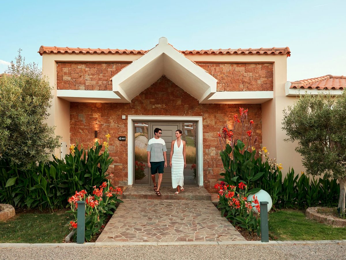 The front of a villa faced in red-brown stone with a terracotta roof and a grey front door, a stone path leading to it between beds of red and yellow canna lilies.