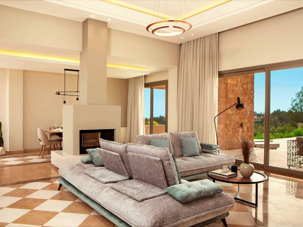 An open-plan villa living room with two long grey sofas set around a central fireplace, a dining table behind and full-height windows onto a terrace.