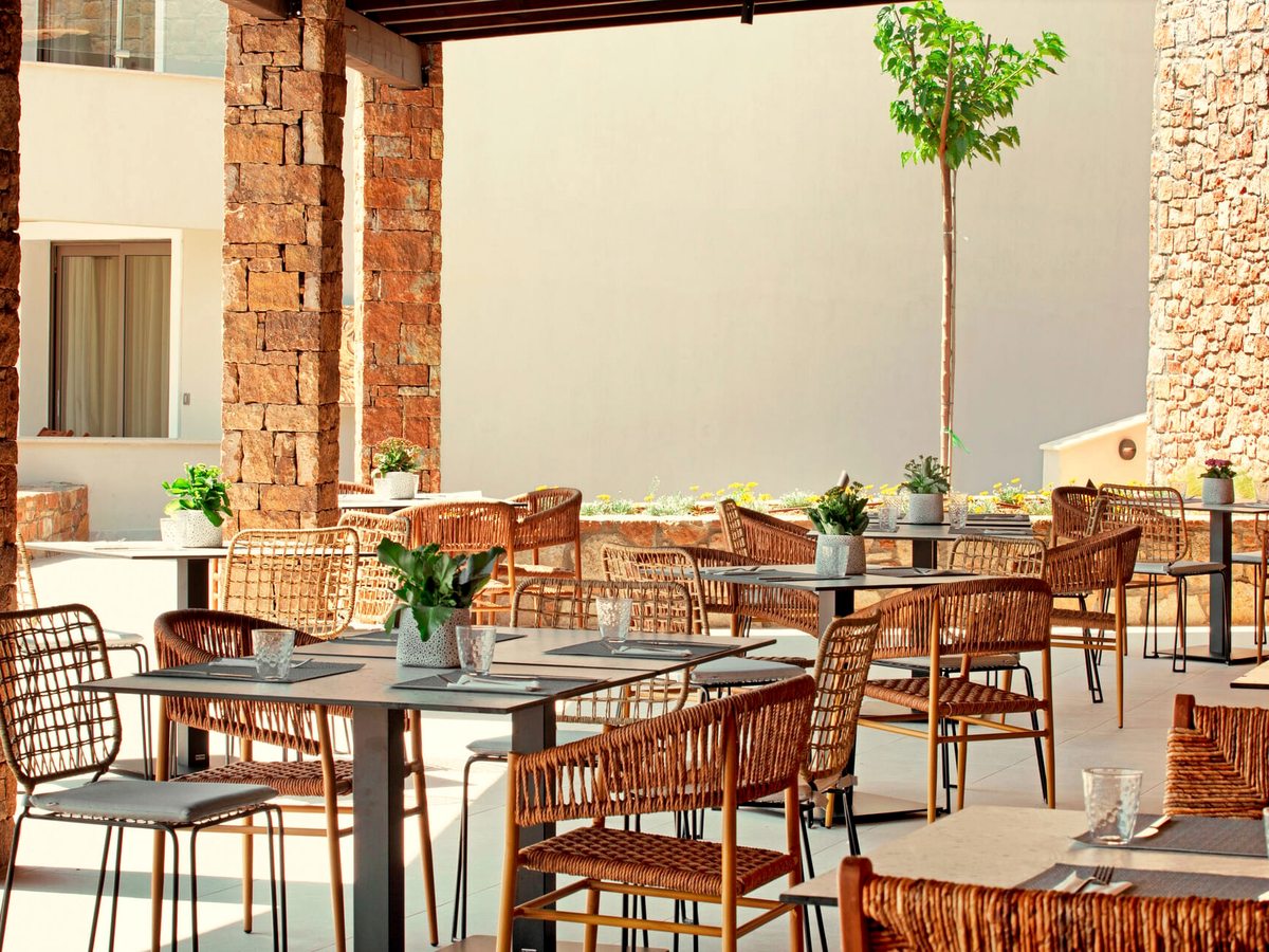 An outdoor dining area under a canopy, with woven wicker chairs at grey tables between rough stone columns and a young tree in a planted bed.