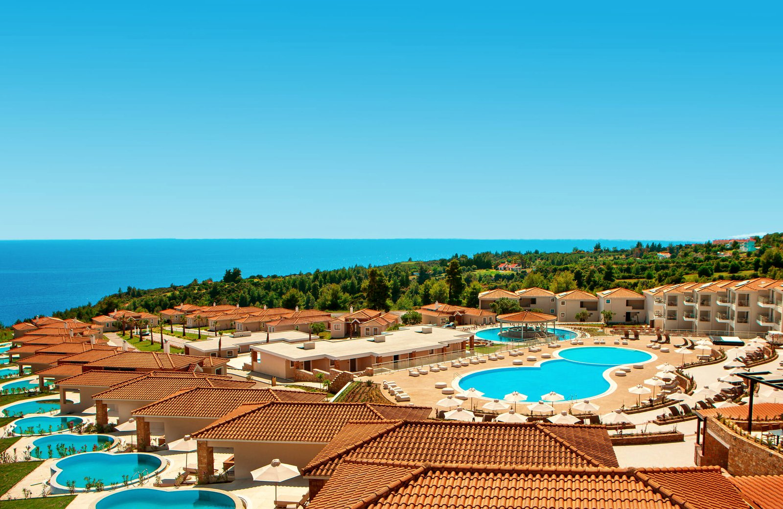 Zeus Eleva Ajul from above: terracotta-roofed bungalows and villas with small private pools stepping down a wooded slope towards two large curved pools, with the Aegean beyond.